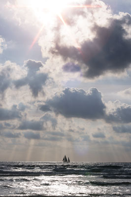 En Donostia-San Sebastián, el sol se oculta tras nubes, pintando el mar y la costa, mientras un velero navega serenamente.