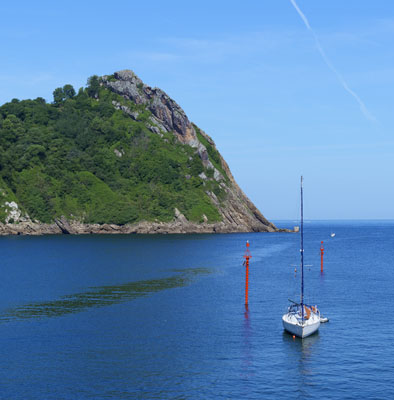 Un elegante velero fondeado descansa en las aguas tranquilas de la bahía de Pasaia. Al fondo, el majestuoso faro de la Plata vigila la costa de Euskadi entre acantilados verdes.